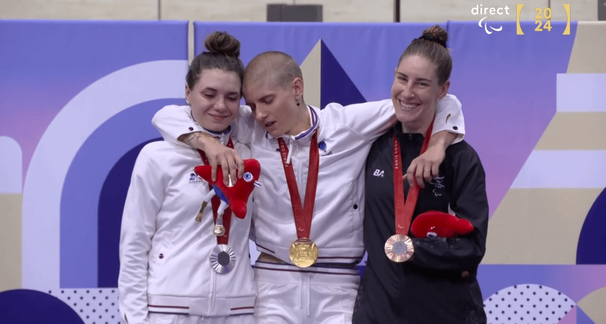 What a shot! Silver and bronze medalists help Marie Patouillet, gold medalist, standing up during her anthem