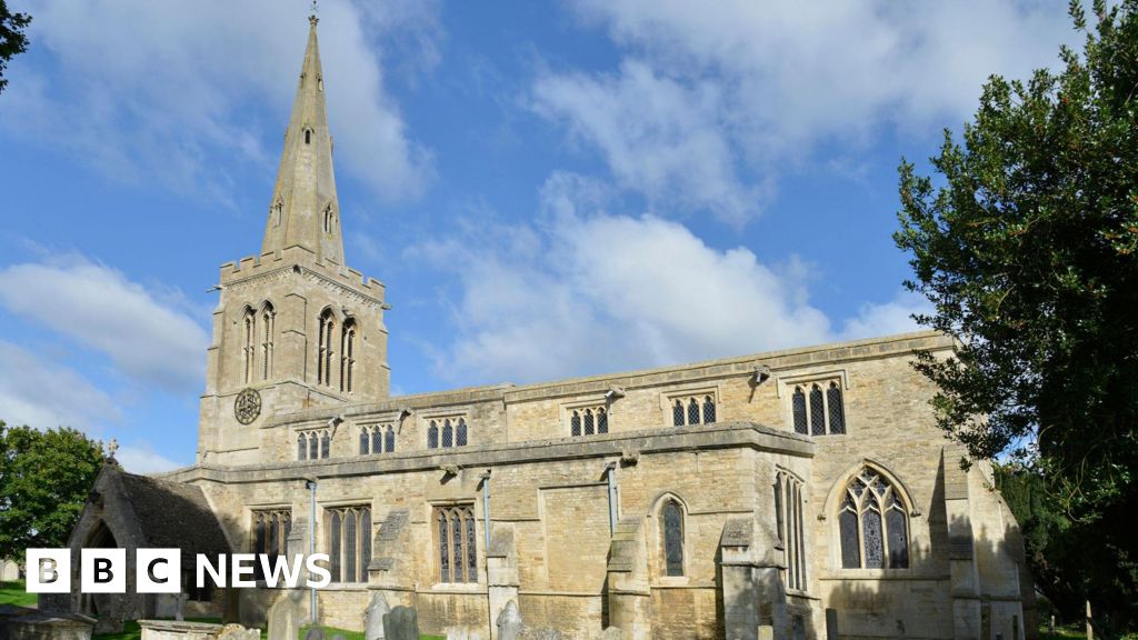 A stone coloured church with a spire on the left hand side. There is a tree on the right and wispy white clouds in the sky.