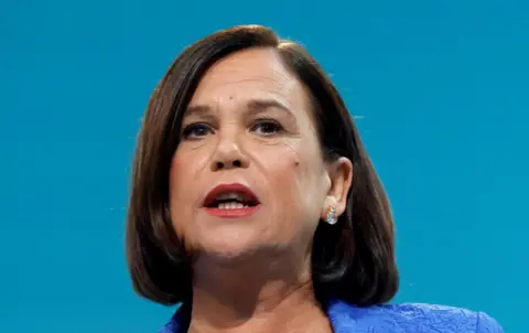 PA Mary Lou McDonald pictured making a speech. She is wearing a blue top and against a light blue backdrop. She has a diamond earring in her left ear