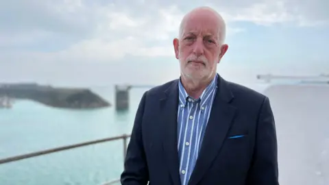 BBC Steve Luce, wearing a dark blazer and a blue and white striped shirt, stood in front of the sea while on a pier in Jersey.