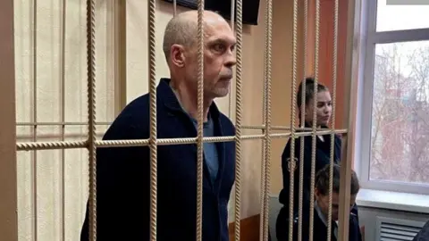 Novosibirsk courts administration Andrey Perlov standing behind bars in court, wearing a dark blue top, there are two guards nearby