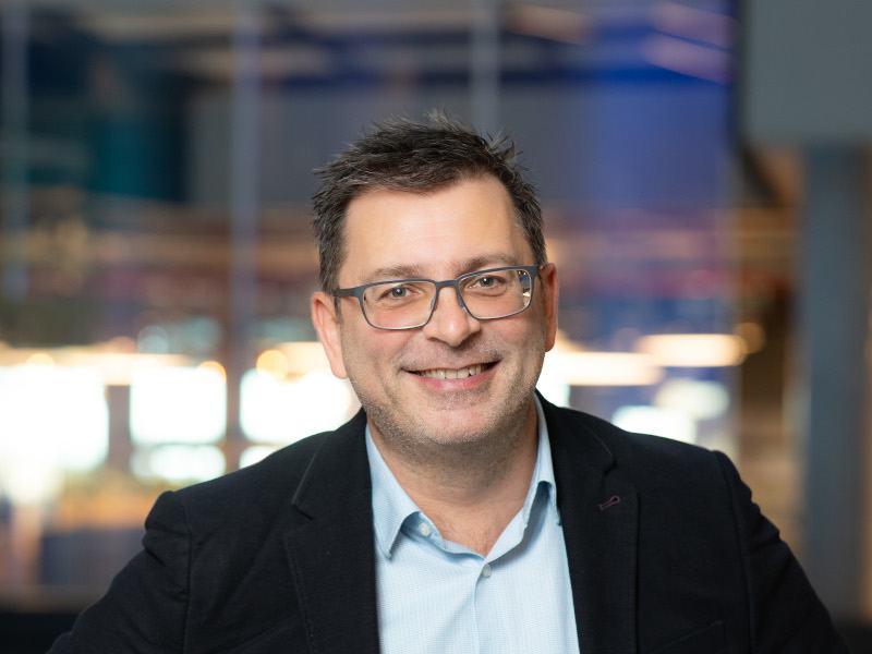 A headshot of a man wearing a black suit jacket, a blue shirt and glasses. He is smiling at the camera.