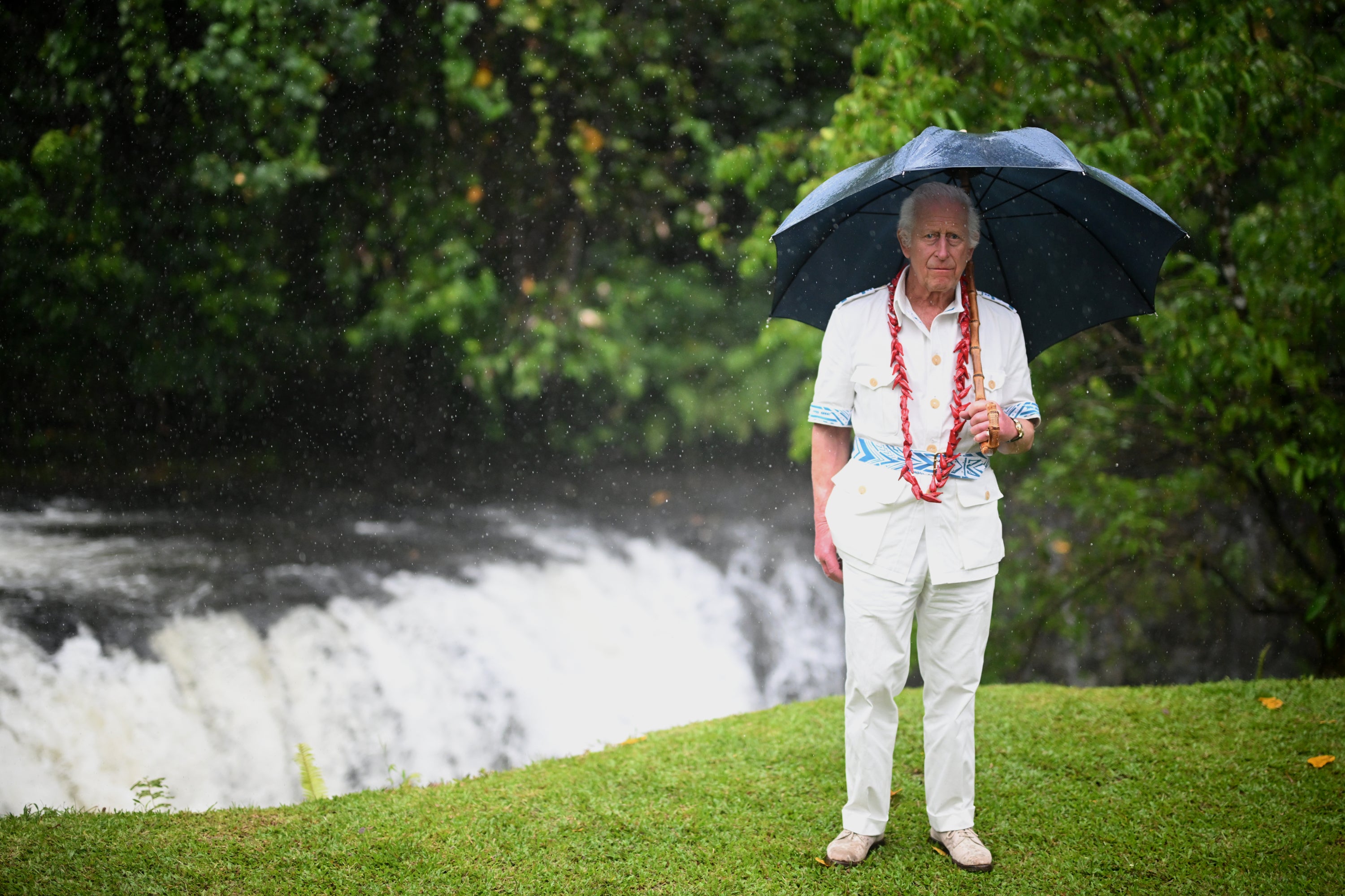 King Charles stands next to a waterfall as he uses an umbrella to shelter from the rain during his visit to O Le Pupu’Pue National Park