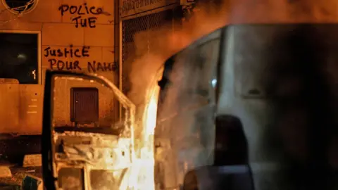 AFP A van burns in front of a building with graffiti reading in French 'Police kill' and 'Justice for Nahel' in Nanterre, France, a day after the killing of 17-year-old Nahel Merzouk - 28 June 2023