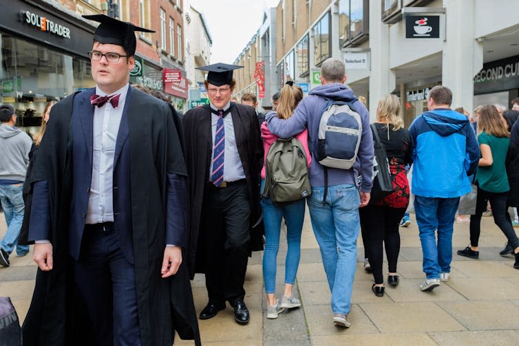 People wearing university graduation gowns walking in one direction and people in ordinary clothes walking in the other.