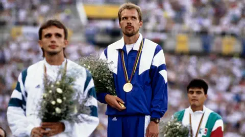 Getty Images Andrey Perlov on the podium at the Olympic Stadium in Barcelona in 1992, wearing a blue and white tracksuit with a gold medal round his neck, holding a bunch of flowers -  on his left and right are the competitors who came second and third in the race