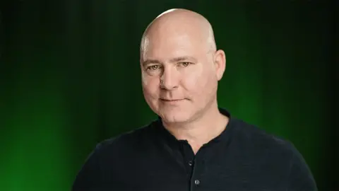 Doug Cockle The voice over actor, Doug Cockle, who is a bald man - smirking with a black buttoned top in front of a black and green background. 