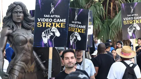 Getty Images Members of SAG-AFTRA striking at WB Studios. They're holding placards saying "SAG-AFTRA Video game strike" with a hand holding a games controller - in front of a wonderwoman statue. 