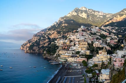 Blick auf Positano, dessen bunte Häuser sich an einen begrünten Hang schmiegen, darunter ein dunkler Strand und das Mittelmeer