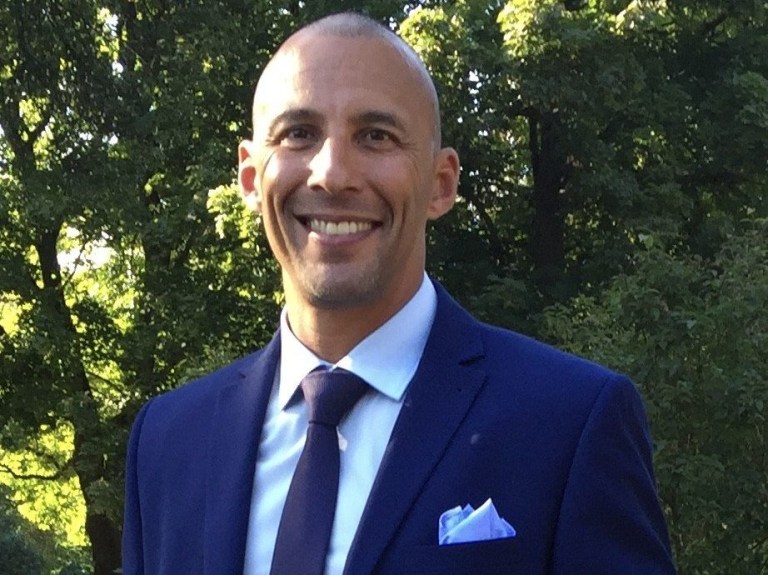 Anthony Sullivan wearing a blue suit, standing in front of some trees