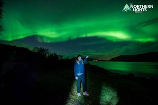 a woman stands in front of a green sky filled with auroras and holds out her hand to point 