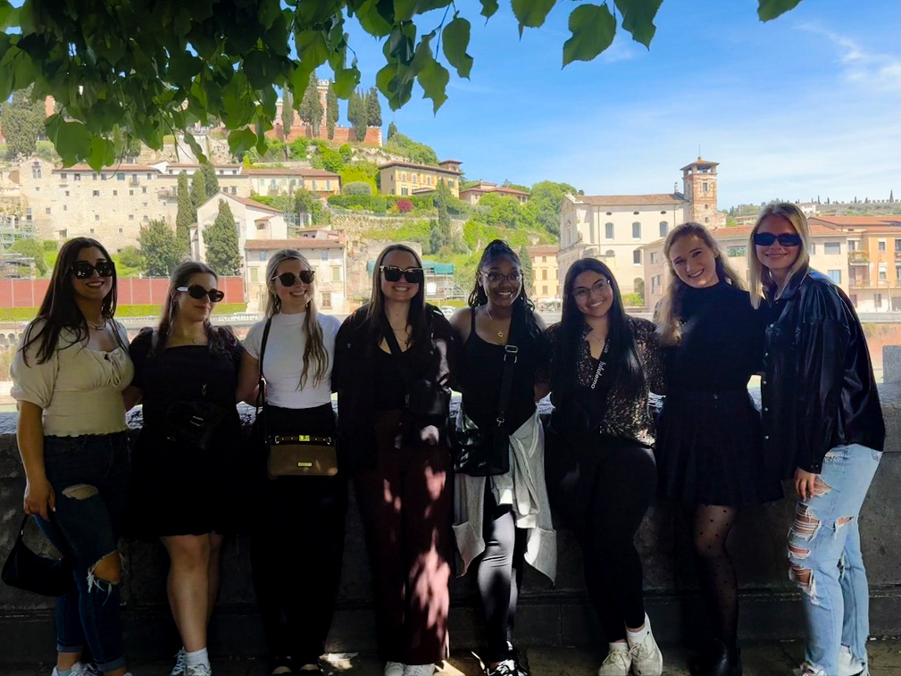 A group of female students in casual attire stand in front of a picturesque Italian village smiling at the camera on a sunny day.