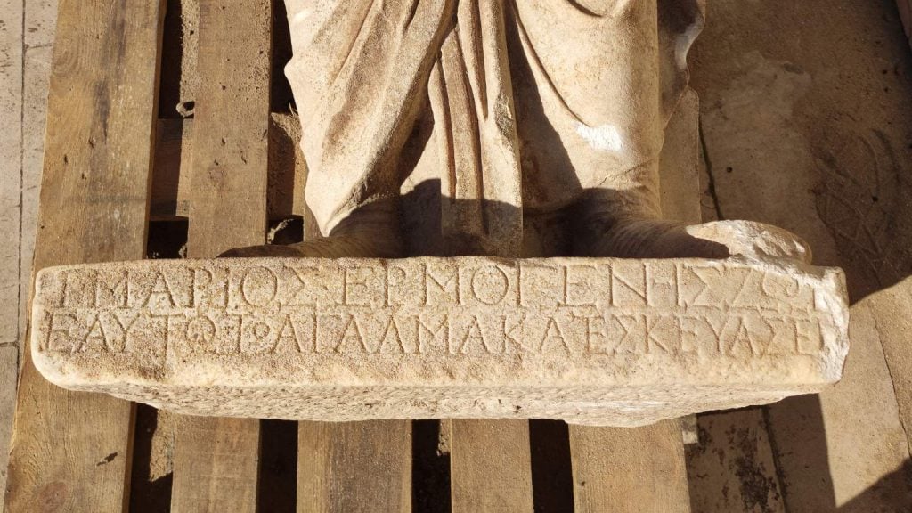 A close-up shot focusing on the base of the stone statue, showing a Greek inscription carved into the stone. The letters are slightly eroded but still legible, providing possible clues to the statue's identity or origin.