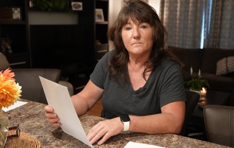 A woman sits at a table in a living room, holding a piece of paper. She doesn't look happy. 