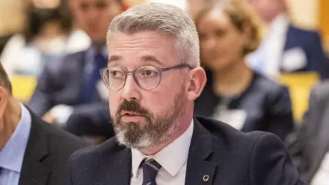 PA Media Niall Ó Donnghaile, pictured from the neck up, wearing a suit and white shirt. He has grey hair and is wearing glasses.