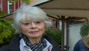 Diana Parkes has short white hair and is wearing a black coat, a pink jumper and a white, buttoned-up shirt and a black scarf. Hetti Barkworth-Nanton is stood next to her. She is smiling and has shoulder length blonde hair. She is wearing a denim jacket and black shirt and a gold necklace around her neck