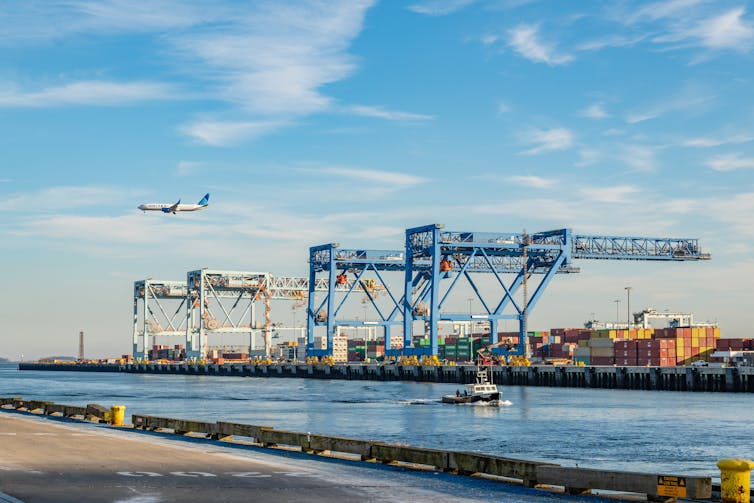 Large loading cranes in harbour ready to load shipping containers onto ships with commercial airplane flying overhead.