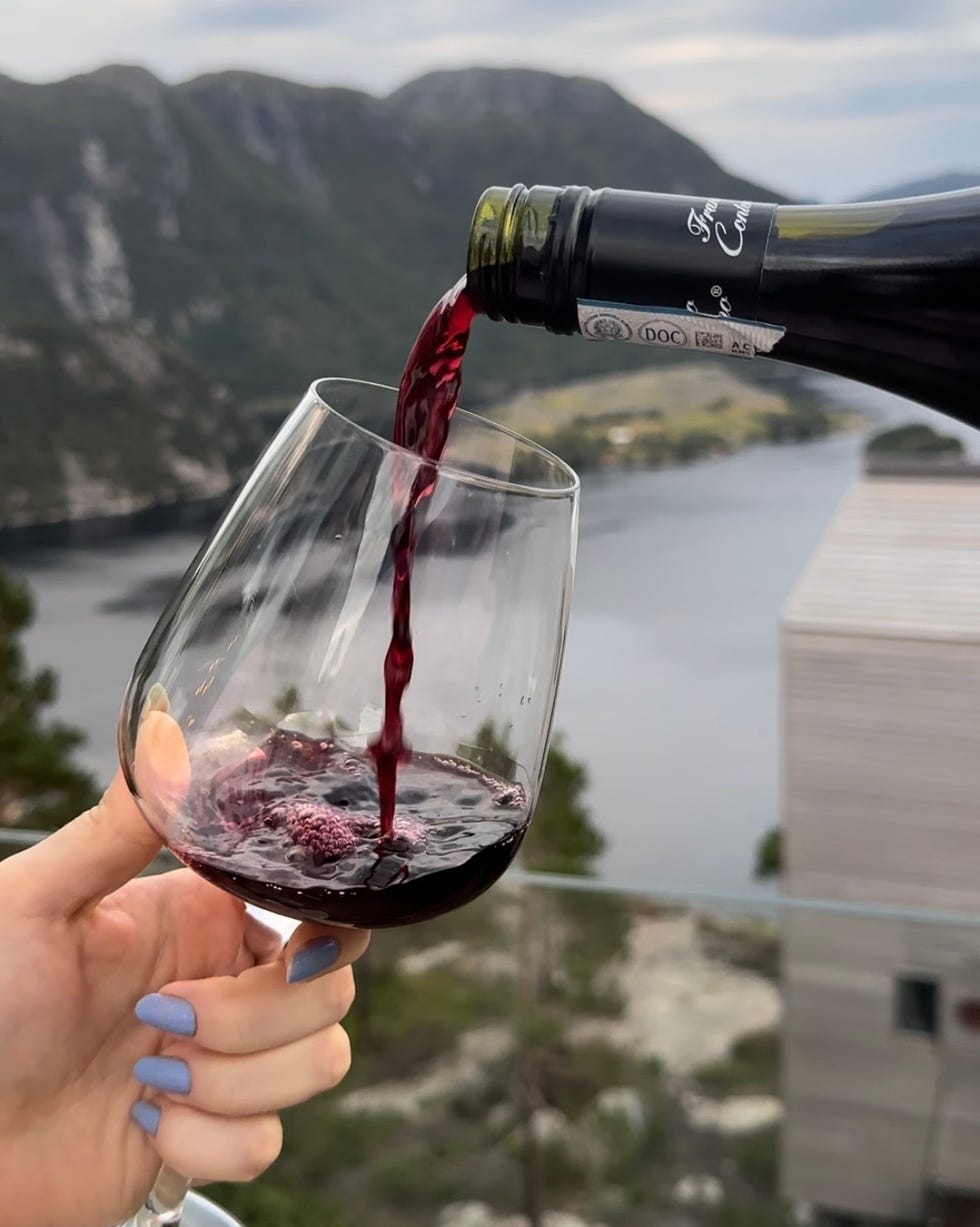 a hand is holding a wine glass as red wine is being poured from a bottle into it the glass is transparent, showing the flowing liquid, while a scenic view featuring mountains and a body of water is visible in the background the atmosphere suggests relaxation and enjoyment