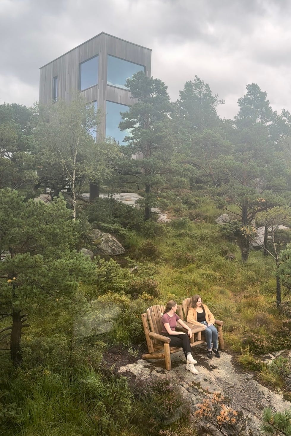 the scene features two people seated on rustic wooden chairs set on a rocky area, surrounded by lush greenery and scattered trees