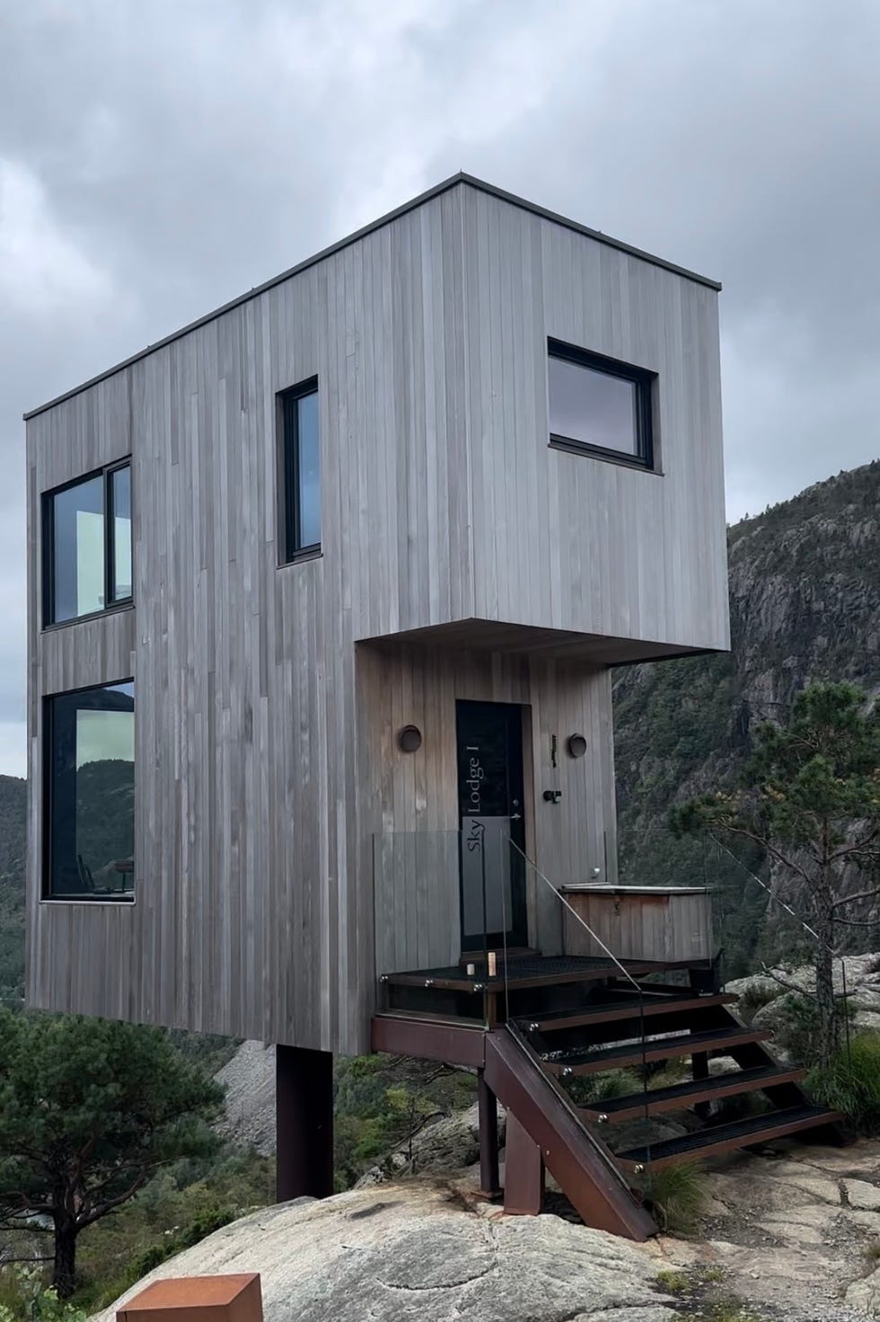 a contemporary cabin with a wooden exterior stands on a rocky landscape, featuring large windows that offer panoramic views the structure is elevated on stilts and includes a set of wooden steps leading to the entrance a small sign indicates 'sky lodge i' next to the cabin, while surrounding greenery and mountains are visible in the background under a cloudy sky