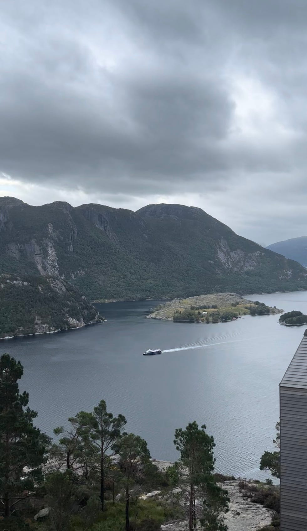 a tranquil fjord is framed by rugged mountains and a lush island a boat is seen gliding across the water, leaving a white wake the sky is overcast, with dark clouds suggesting a potential for rain in the foreground, there are trees and rocky terrain, adding to the natural beauty of the landscape