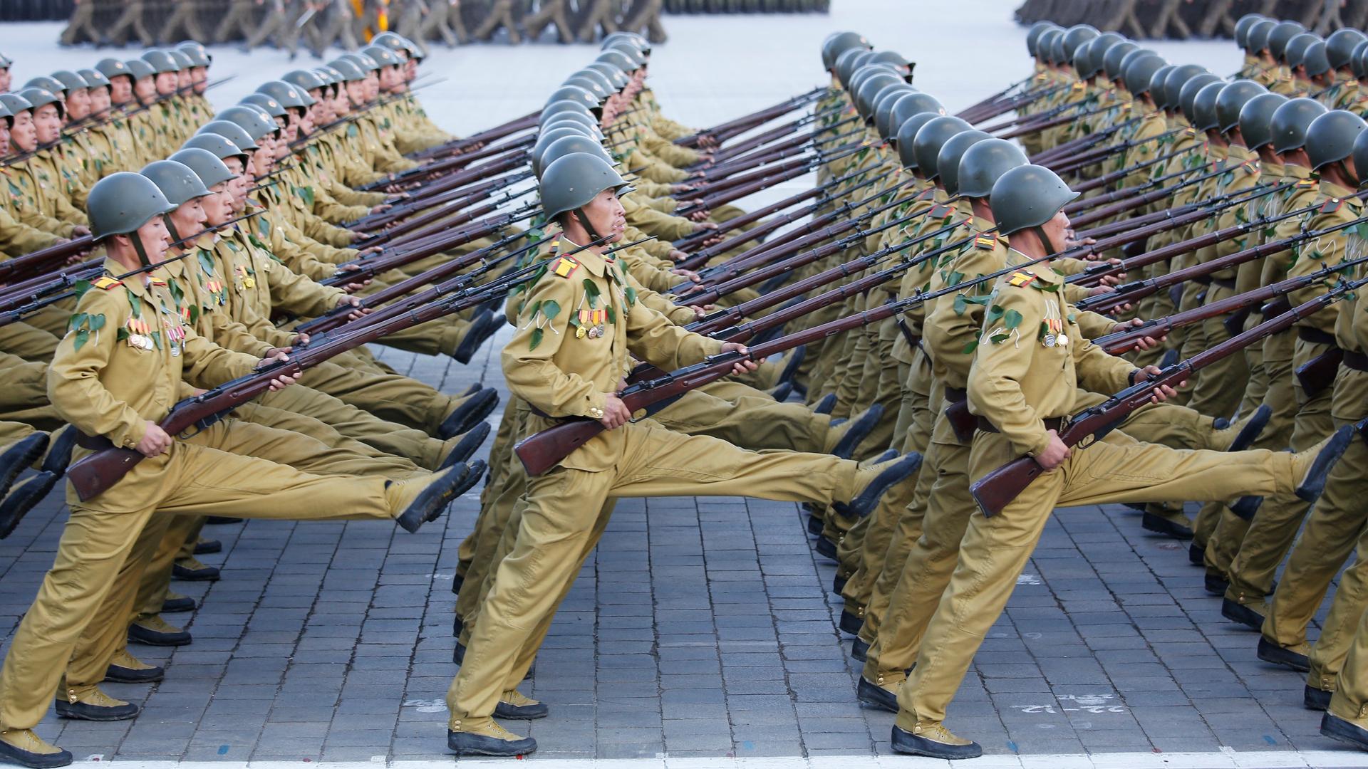 Die Soldaten gehen in mehreren Reihen hintereinander im Stechschritt über einen Platz. Sie tragen Gewehre. 