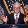 This photo shows Federal Reserve Chair Jerome Powell standing behind a lectern that has two microphones. He's wearing a suit and glasses. American flags and also flags with the Federal Reserve seal are in the background.