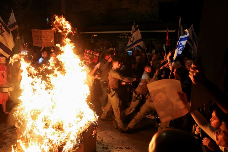 police clash with protestors waving israeli flags next to a fire