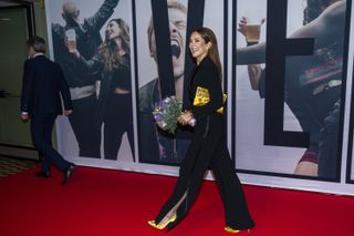 Queen Mary walking down the red carpet wearing a black jumpsuit with yellow shoes and holding flowers