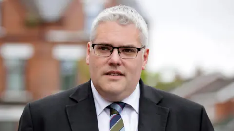 PA Media A man with short grey hair looks off to the side of the camera. He is wearing glasses, a dark suit jacket, white shirt and blue and green stripe tie. The background is blurred, but shows the outline of red brick buildings. 