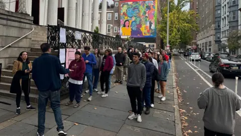 Two lines of people on a sidewalk, most of hem wearing dark sweats 