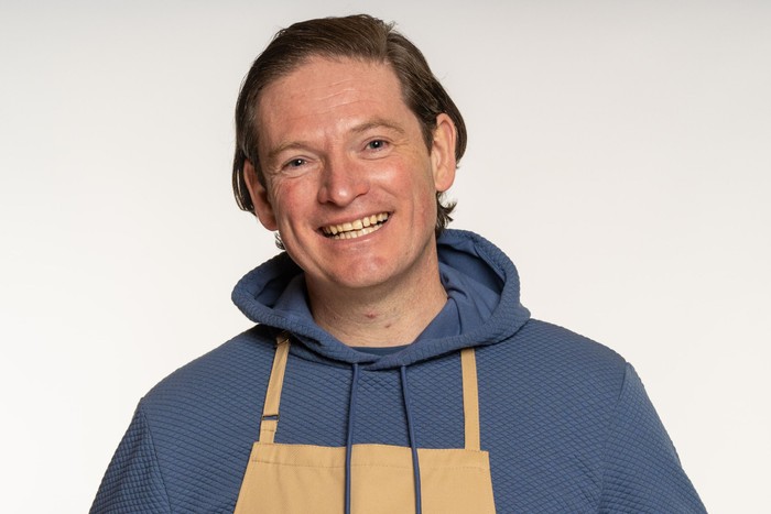 Andy wearing a blue hoodie and brown apron, smiling ahead with his hair slicked back.