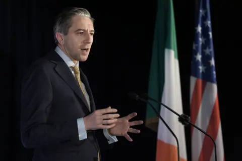 PA Simon Harris in grey suit jacket, blue shirt and gold tie, stood in front of a USA and Ireland flags, speaking into microphones at press event