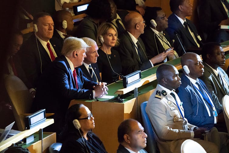 Two old men in suits confer, surrounded by other people in suits.