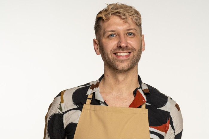 John wearing a floral shirt and brown apron, smiling ahead.