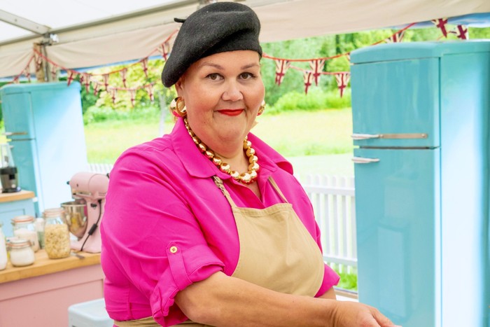 Nelly wearing a pink shirt, brown apron and a black beret - smiling ahead.